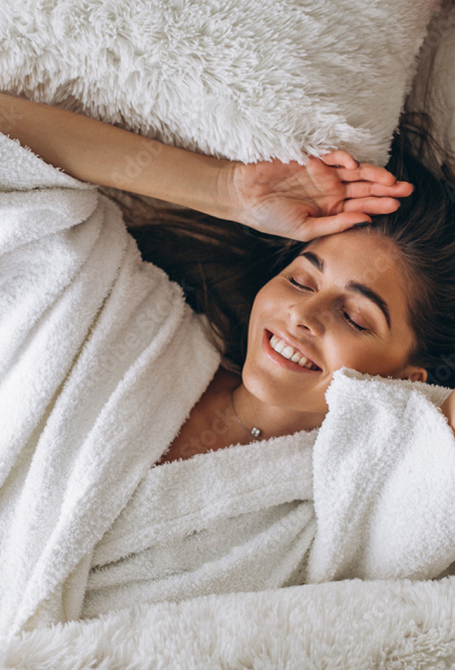 Eine lächelnde Frau entspannt in einem weißen Bademantel auf flauschigen Kissen.