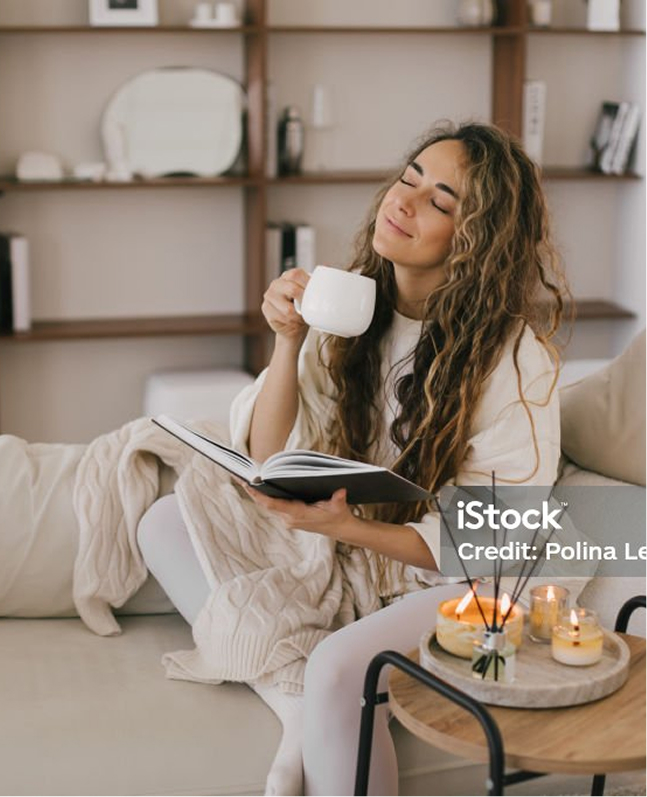 Eine junge Frau genießt entspannt ein Heißgetränk auf dem Sofa, im Vordergrund brennen Kerzen.