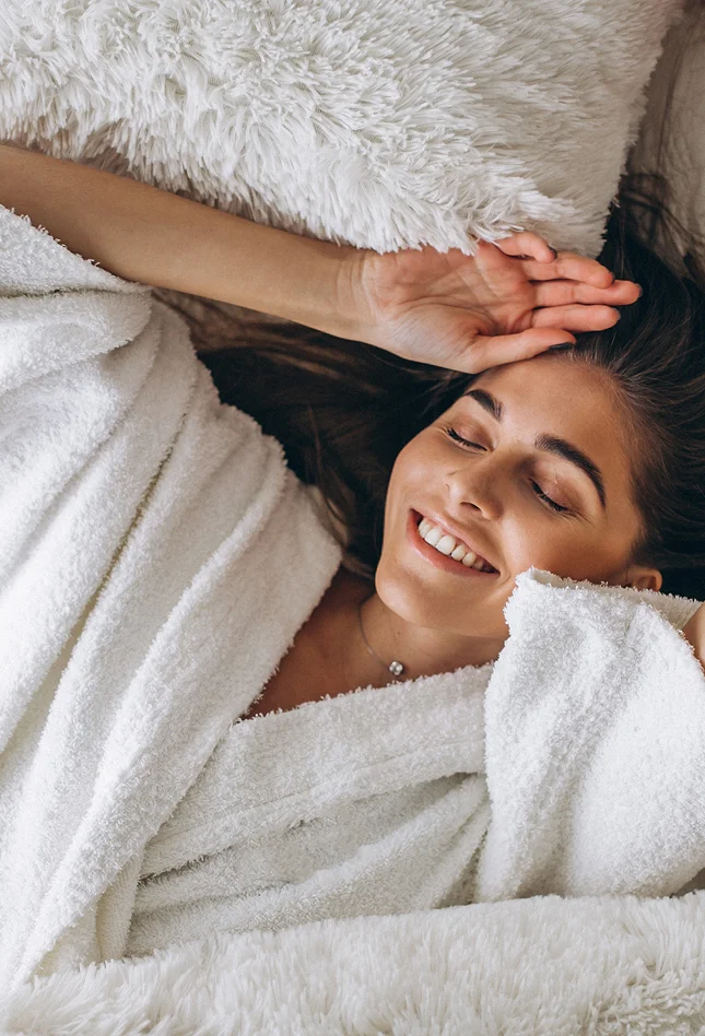 Eine lächelnde Frau entspannt in einem weißen Bademantel auf flauschigen Kissen.
